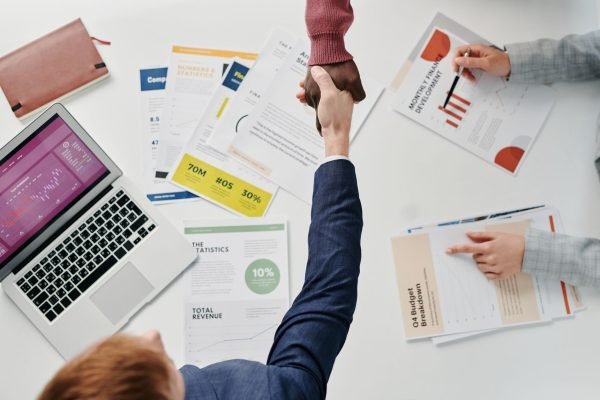Business professionals engaging in a handshake over a desk filled with financial documents and a laptop. — Essential Skills f