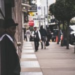 A man in a suit and hat leans against a city building, observing pedestrians. — Reliable Nangs Delivery for Every Occasion
