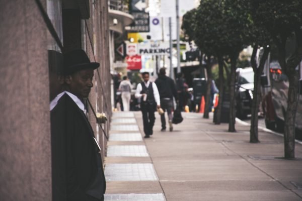 A man in a suit and hat leans against a city building, observing pedestrians. — Reliable Nangs Delivery for Every Occasion