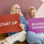 Two women on a sofa holding signs promoting startup and business women. — The Unsung Heroines of Indian Households: A Tribute